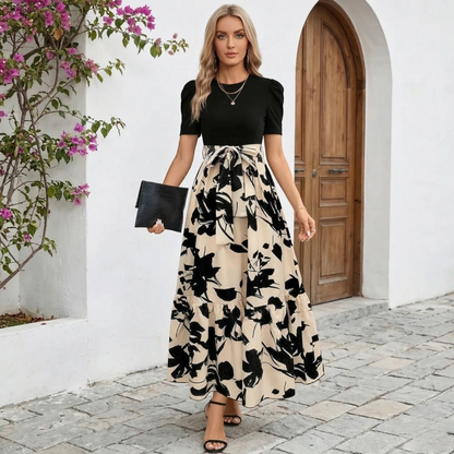 Olive-Black Leaf - A woman wearing a mock two-piece maxi dress with a fitted black short-sleeve bodice and a high-waisted cream skirt featuring a bold black abstract floral print, styled with a self-tie waist and tiered hem.
