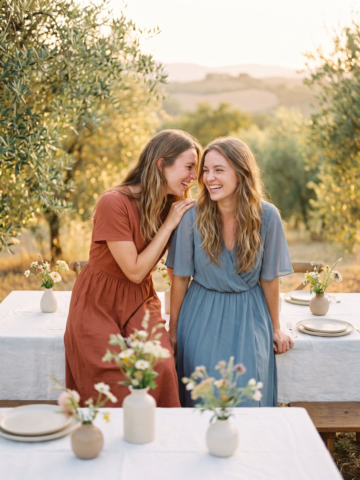 Two friends laughing at an outdoor lunch table in an olive grove, in terracotta and dusty blue midi dresses